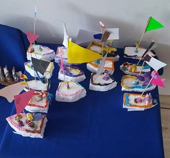 Ważenie przedmiotów i budowa łódek. Na ciemno niebieskim obrusie widać kilkanaście styropianowych łódek z trójkątnymi, papierowymi, kolorowymi żagielkami.