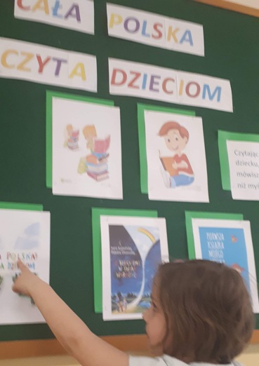 Na tle zielonej tablicy widoczny jest napis „CAŁA POLSKA CZYTA DZIECIOM" oraz obrazki z dziećmi czytającymi książki. Przed tablicą stoi dziewczynka wskazująca palcem na jeden z obrazków.