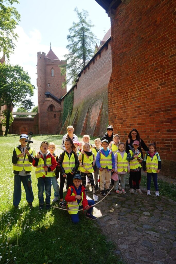 Grupa dzieci w kamizelkach odblaskowych na tle murów zamku w Malborku