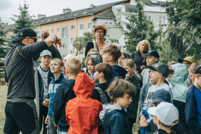Grupa dzieci słucha pana prowadzącego zajęcia na tle drzew i bloku.